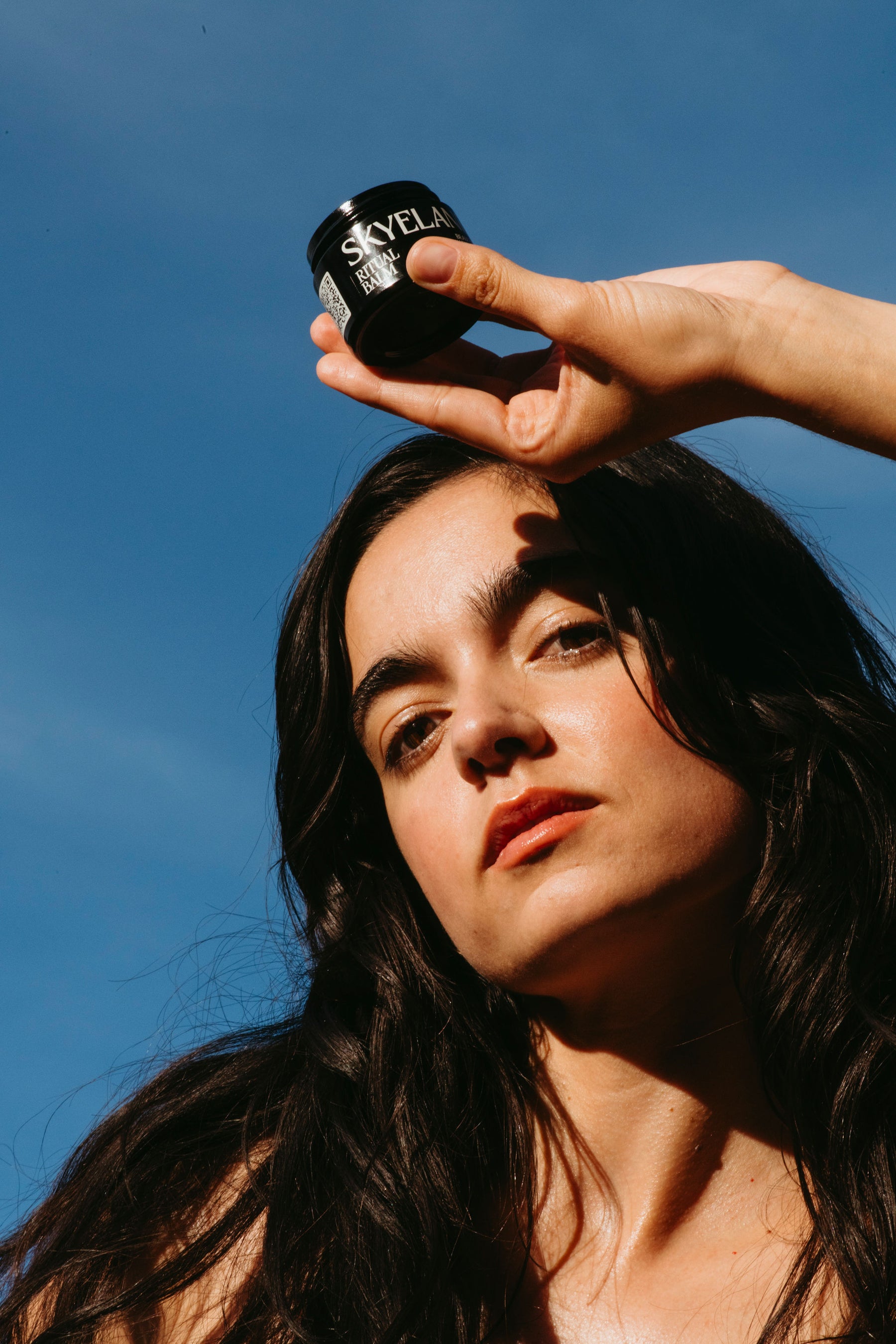 A woman outdoors under a clear blue sky holds a small black jar labeled SKYELAND above her head with her hand. The product is waterless botanical skincare called ritual balm, shown in a lifestyle setting with strong natural sunlight and shadows.